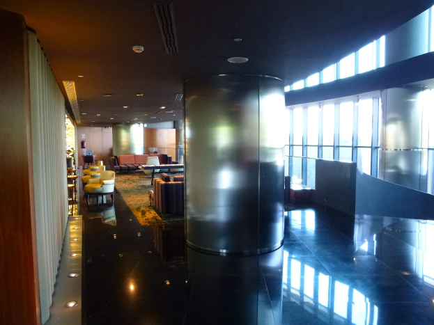 Eine geräumige First-Class-Lounge am Hong Kong International Airport mit großen Fenstern, hohen Decken mit elektrischen Leuchten, Sitzmöglichkeiten einschließlich Stühle und Sofas, Tischen, einem Teppichboden und einer zentralen Säule.