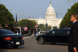 Trauerzug mit Menschen in Mützen und Fahnen vor dem Kapitolsgebäude, Fahrzeuge, Laternenpfähle, Bäume und Gebäude unter einem klaren Himmel.