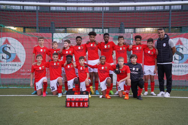 Eine Gruppe junger Männer in roten T-Shirts und schwarzen Shorts steht auf einem Fußballfeld neben einer Kiste roter Gegenstände, mit Schildern im Hintergrund, die "FC Rot-Schwarz Eglaute - Champions League" lesen.