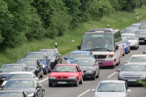 Ein Stau auf einer Autobahn mit vielen Autos und einem Van, Menschen sind in den Fahrzeugen zu sehen, mit Bäumen und Gras im Hintergrund.