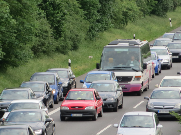 Ein Stau auf einer Autobahn mit vielen Autos und einem Van, Menschen sind in den Fahrzeugen zu sehen, mit Bäumen und Gras im Hintergrund.