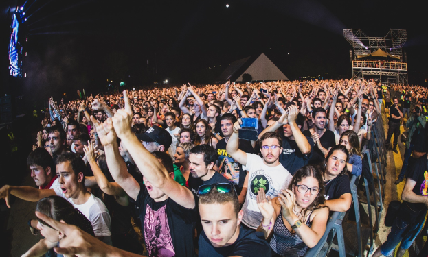 Eine große Menge bei einem Musikfestival mit erhobenen Händen, einige sitzen auf Stühlen und andere stehen, begrenzt von einem Metallzaun auf der rechten Seite, mit Gebäuden und Lichtern im Hintergrund unter einem dunklen Himmel.