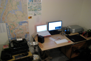 Ein Raum mit einem Tisch, auf dem ein Laptop, ein Lautsprecher, ein Monitor, eine Tastatur, ein Ball, eine Maus, ein Scanner und Papiere liegen; ein weiterer Tisch mit Elektronikgeräten im Vordergrund; und eine Wand mit Charts und Postern im Hintergrund.