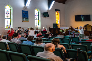 Eine Gruppe von Menschen sitzt auf Stühlen in einer Kirche, ein Mann steht vorne mit einem Mikrofon, umgeben von Musikinstrumenten, einem Tisch mit einer Blumenvase, einem Lautsprecher, einer Wand mit Text und Fenstern, einem Fernseher und Deckenleuchten.