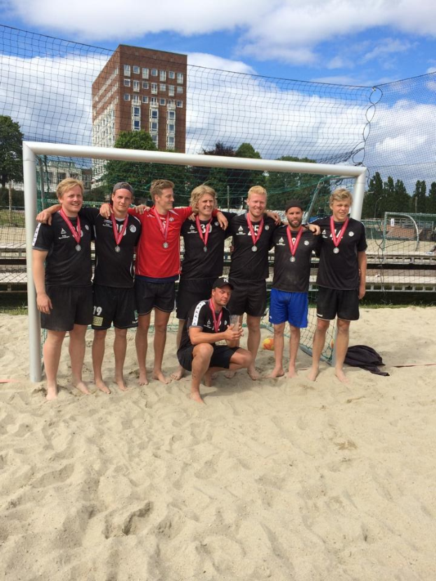 Mannergruppe in sportlicher Kleidung auf einem sandigen Strand, mit Medaillen und umgeben von einem Netz, mit Bäumen, Gebäuden und einem bewölkten Himmel im Hintergrund.