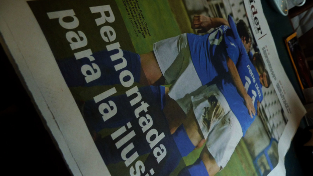 Eine Zeitung auf einem Tisch zeigt drei Männer in blauen T-Shirts, möglicherweise beim Fußballspielen, mit Text unten auf dem Bild.