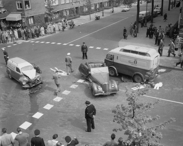 Eine Schwarz-Weiß-Szene eines Autounfalls an einer Straßenkreuzung, mit mehreren Fahrzeugen einschließlich eines Lieferwagens, einer Gruppe von Menschen drumherum und Gebäuden mit Fenstern, Bäumen, Pfählen und Texttafeln im Hintergrund.