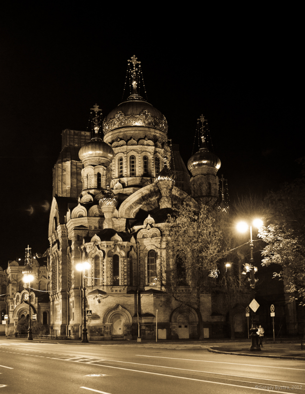 Eine Straße mit weißen Linien im Vordergrund, mit Laternenmasten und Gebäuden im dunklen Hintergrund.