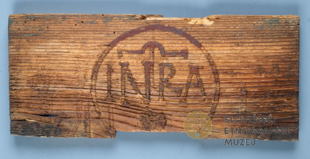 Ein Stück Holz mit der eingravierten Aufschrift 'NRA', mit einem Wasserzeichen und vor einem blauen Hintergrund.