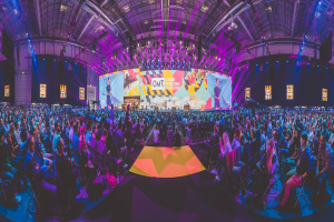 Eine große Menschenmenge auf Stühlen sitzend, die eine hell erleuchtete Bühne mit mehreren stehenden Individuen, wahrscheinlich auf einem Online-Marketing-Rockstars-Event, umgeben.