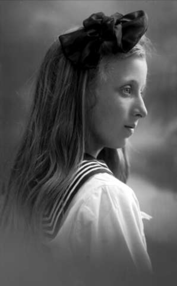 Ein junges Mädchen in einer Matrosenuniform mit einer Schleife auf dem Kopf, ihr Gesicht weich beleuchtet vor einem unscharfen, träumerischen Hintergrund.