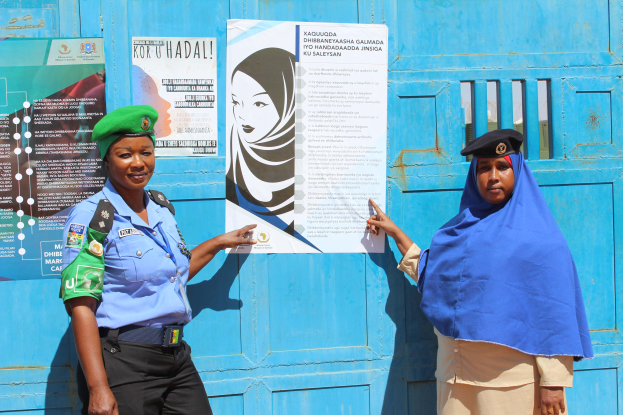 Zwei Frauen stehen nebeneinander vor einer blauen Tür, die mit Plakaten bedeckt ist. Eine trägt ein blaues Hemd, schwarze Hosen und eine grüne Mütze, die andere hält ein Papier in der Hand, was darauf hindeutet, dass sie Teil einer Polizeitruppe sind.