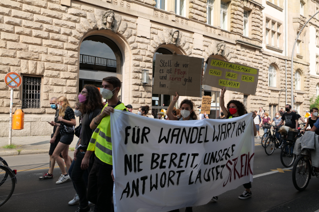 Eine Gruppe von Menschen marschiert auf einer Straße in Berlin, hält Plakate und Banner hoch und fährt dabei Fahrräder, vor einem Gebäude mit Bögen, Säulen, Skulpturen und Bäumen.