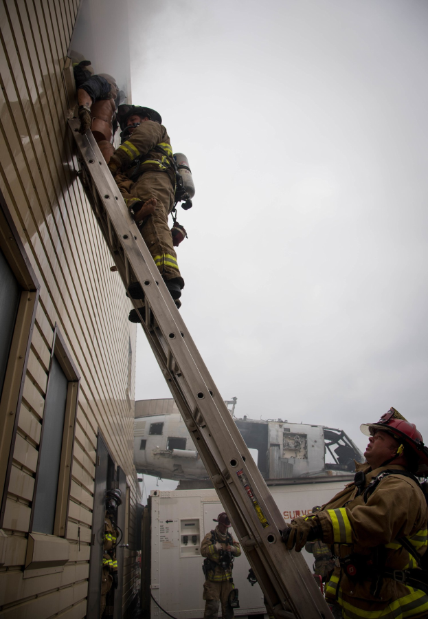 Feuerwehrleute mit Helmen, die ein Gebäude mit einer Leiter hochklettern, mit einem Fahrzeug im Hintergrund.