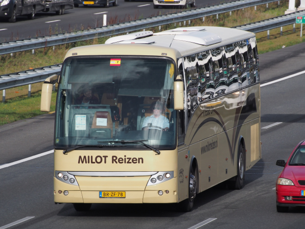 Ein Minibus mit sichtbaren Insassen auf einer Autobahn neben einem roten Auto fahrend, mit Geländern und Gras am Straßenrand und Texttafeln und Säulen im Hintergrund.