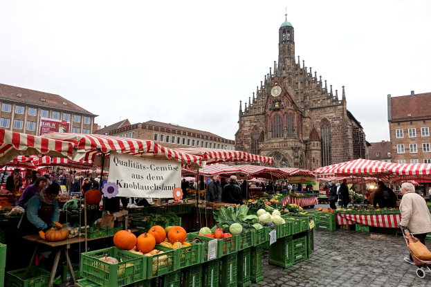 Ein belebter Markt in Nürnberg, Deutschland, mit Ständen, die verschiedene Früchte und Gemüse anbieten, Menschen mit Taschen und Zelten im Hintergrund sowie Gebäude und einen Uhrenturm.