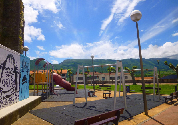 Spielplatz mit Schaukeln, einer Rutsche, Bänken, Pfosten, Laternen, einer bemalten Wand, Gras, Bäumen, Gebäuden, Hügeln und einem bewölkten Himmel im Hintergrund.