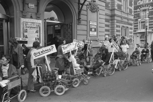 Eine Gruppe von Menschen im Rollstuhl mit Schildern an der Straße, im Hintergrund Gebäude und ein Laternenpfahl auf der rechten Seite, in Schwarz-Weiß.