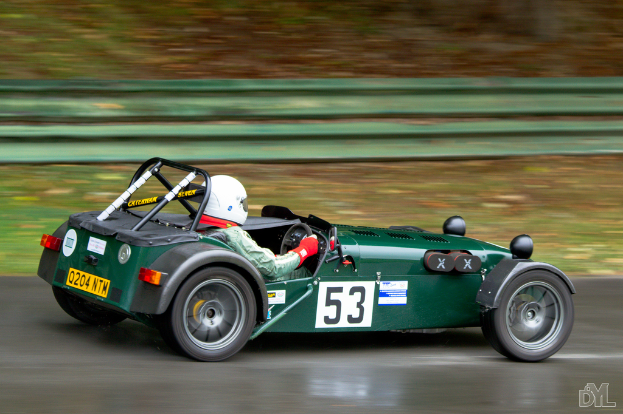 Eine Person mit Helm fährt ein Auto mit Logo unten rechts, Gras ist im Hintergrund sichtbar.