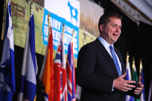 Ein Mann im Anzug und Krawatte steht an einem Rednerpult mit einem Mikrofon, umgeben von Fahnen und einem Bildschirm im Hintergrund, und scheint eine Äußern zu machen.