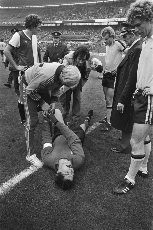 Schwarzes Bild eines Fußballspielers auf dem Boden umgeben von einer Gruppe von Männern, mit einer Zuschauermenge im Hintergrund eines Stadions.