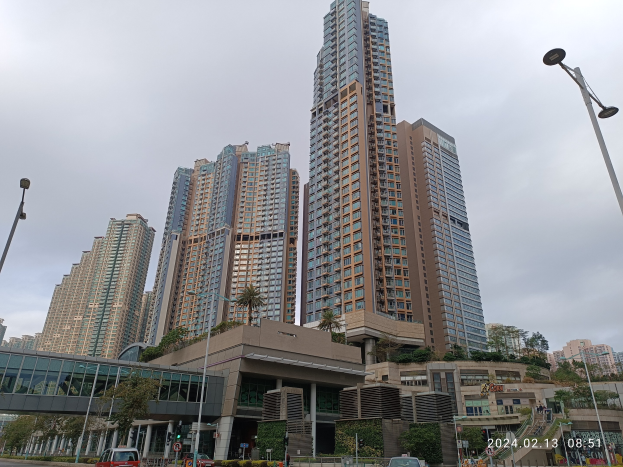 Eine Stadtstraße mit hohen Gebäuden im Hintergrund, Fahrzeugen auf der Straße, Laternen, einer Brücke, Bäumen, Pflanzen und einem bewölkten Himmel, mit einem hohen Wohngebäude im Vordergrund.