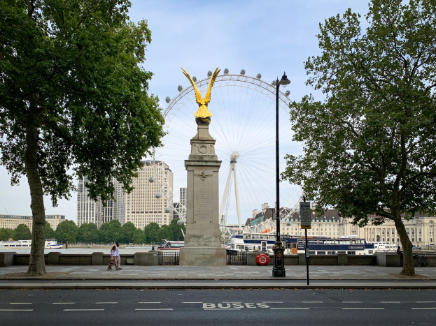 Das London Eye mit einer Statue auf einem Podest im Vordergrund, eine Straße, Bäume, ein Laternenpfahl, Menschen, die spazieren gehen, Schiffe auf dem Wasser, Gebäude und der Himmel.