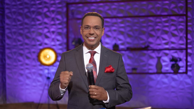 Stephen Colbert, der einen Anzug, eine Krawatte und eine Brille trägt, steht auf einer Bühne und hält ein Mikrofon in der Hand, während sich hinter ihm eine Wand und verschiedene Gegenstände befinden.