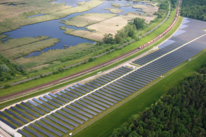 Luftaufnahme eines Solarparks mit Solarpanelen auf einem Feld, umgeben von Bäumen, Gras, Pflanzen und Wasser, mit einem Zug auf einer nahen Bahnstrecke.