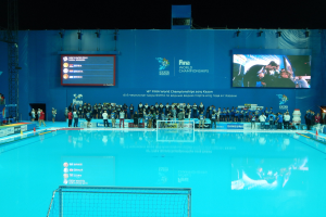 Eine Gruppe von Menschen um ein Hallenbad mit Netz am Boden herumstehend, mit einer Wand im Hintergrund, die "FINA World Championships 2015 Kazan" anzeigt.