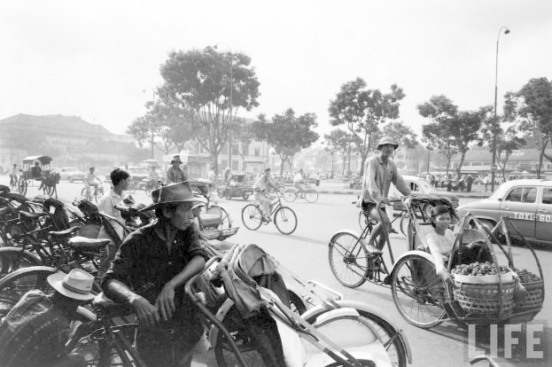Eine vielbefahrene Straße mit verschiedenen Fahrzeugen, darunter Fahrräder und Autos, mit Bäumen und Gebäuden im Hintergrund.