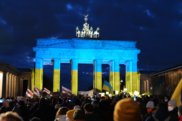 Eine Menschenmenge steht vor dem Brandenburger Tor in Berlin, Deutschland, mit Fahnen und Schildern in den Händen, einem Banner auf der rechten Seite und dem Torbogen, der mit Statuen und Säulen geschmückt ist, unter einem bewölkten Himmel.