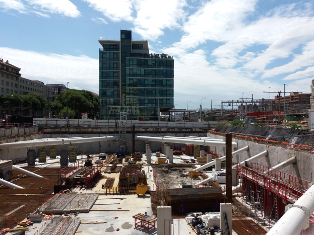 Baustelle mit einem großen Gebäude im Hintergrund, umgeben von Bäumen, Pfählen, Lampen und anderen Gegenständen, unter einem bewölkten Himmel, als Europäischer Gerichtshof identifiziert.