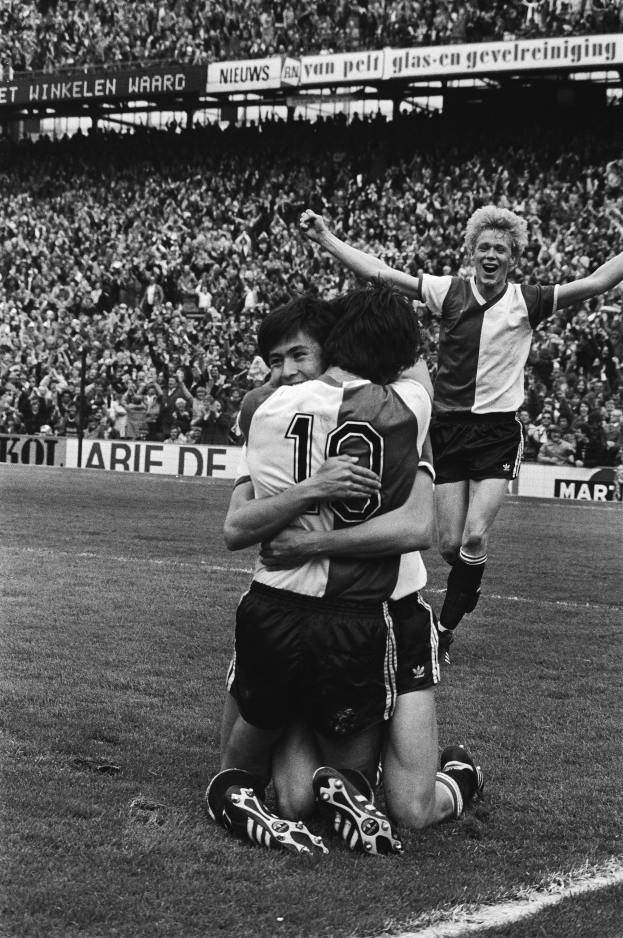 Drei Fußballer umarmen sich auf dem Boden und feiern ein Tor vor einer jubelnden Menge in einer schwarz-weißen Stadionszene.