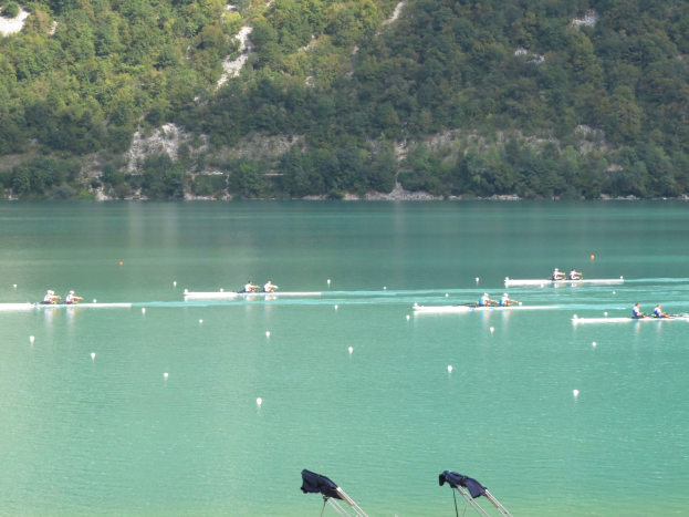 Eine Gruppe von Menschen, die auf einem Gewässer umgeben von Bäumen und einem Hügel rudern, mit Bekleidung-behangenen Tribünen im Hintergrund, die auf einen Ruderwettbewerb hinweisen.