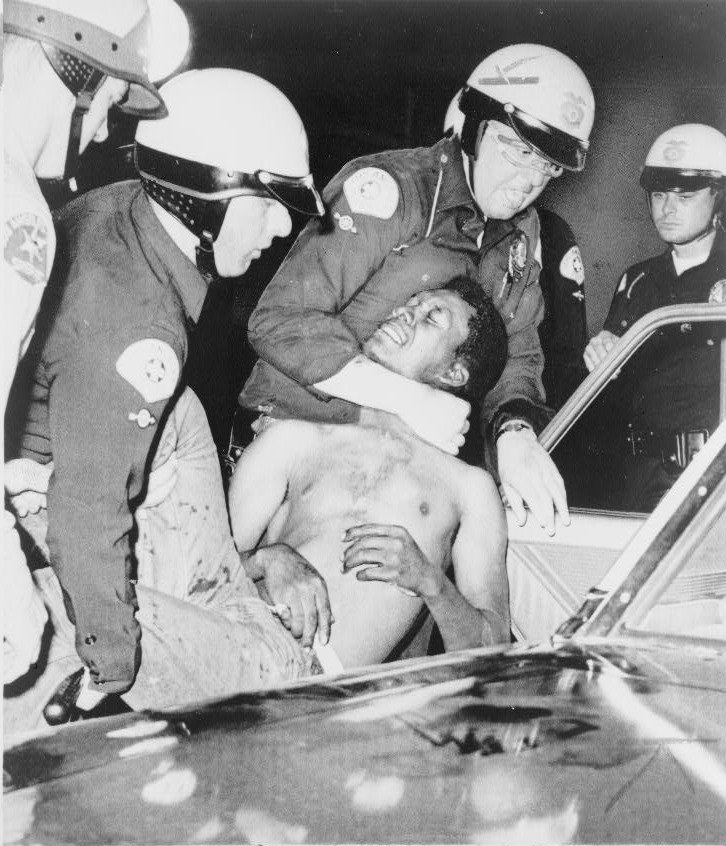 Schwarz-weiß-Foto eines Mannes in Not, der von Polizeibeamten in Helmen begleitet wird.
