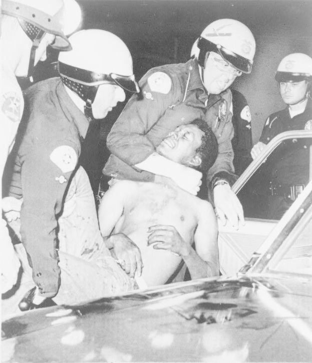 Schwarz-weiß-Foto eines Mannes in Not, der von Polizeibeamten in Helmen begleitet wird.