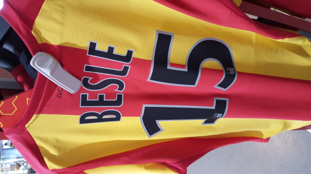 Roter und gelber Fußballtrikot mit Text und Zahlen an einem Ladengestell hängend, mit anderen Gegenständen und dem Hintergrund im Hintergrund.