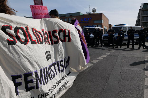 Eine Gruppe von Menschen marschiert mit einem Banner, auf dem 'Solidarität und Feminismus' steht, die Straße entlang, mit parkenden Fahrzeugen, Gebäuden, einer Schüsselantenne und einem klaren blauen Himmel im Hintergrund.