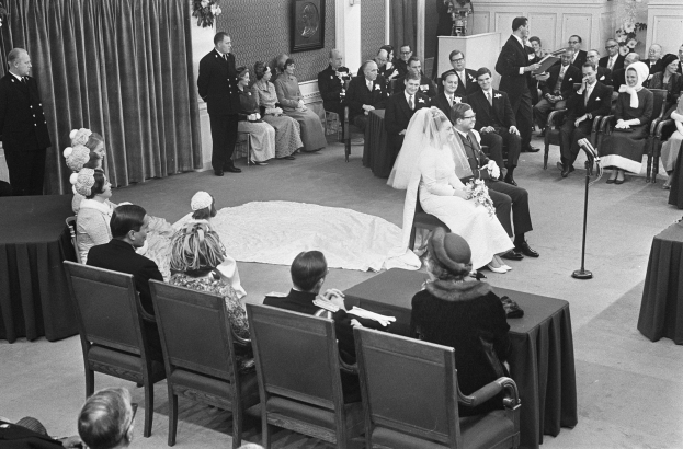 Schwarzes und weißes Foto einer Hochzeitszeremonie mit sitzenden und stehenden Gästen, einem Mann mit Mikrofon, einer Frau mit Blumenstrauß, einem Podium mit Mikrofon auf der rechten Seite, Blumenschmuck und einem Vorhang auf der linken Seite, einem gerahmten Foto an der Wand und einer Tür im Hintergrund.
