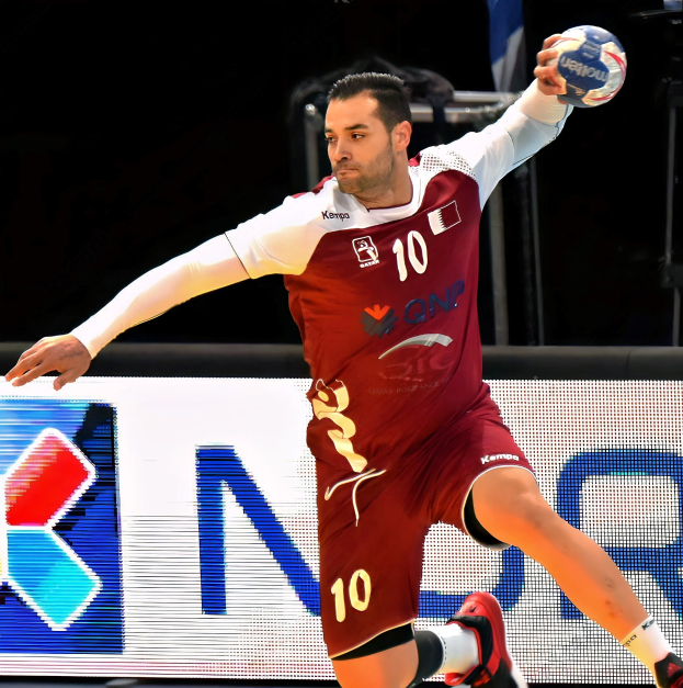Mann in rot-weißer Uniform spielt Handball, hält Ball und hat entschlossene Miene, Arme ausgestreckt, vor "QNP Handball World Cup 2019"-Schild.