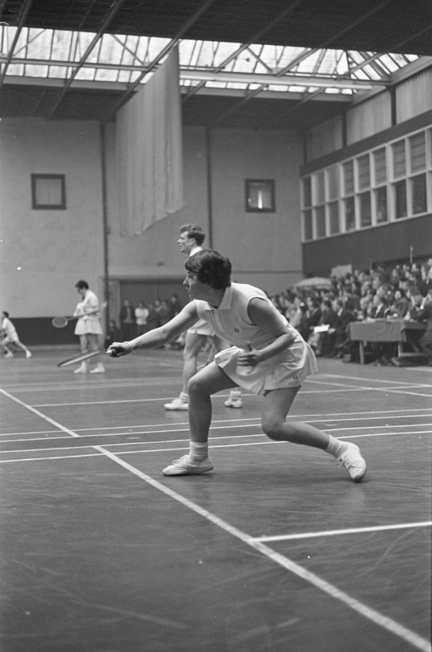 Schwarz-weiß-Foto von zwei Frauen, die Badminton auf einem Court spielen, jede hält einen Schläger, mit Zuschauern rechts und einer Wand mit Fenstern im Hintergrund.