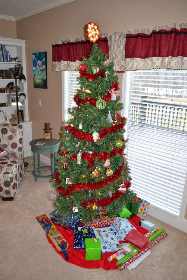 Ein Weihnachtsbaum mit Geschenken davor, eine Couch, ein Tisch, ein Bücherregal, eine Stab-Lampe und ein Bild an der Wand, alles in einem Raum mit Vorhängen an den Fenstern.
