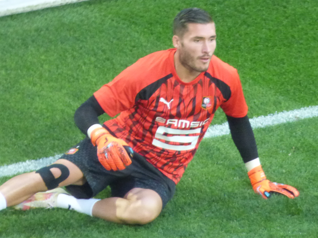 Ein Fußballspieler in rotem T-Shirt, schwarzen Shorts, weißen Socken und orangefarbenen Baseball-Handschuhen sitzt auf dem Boden mit Händen auf den Knien.