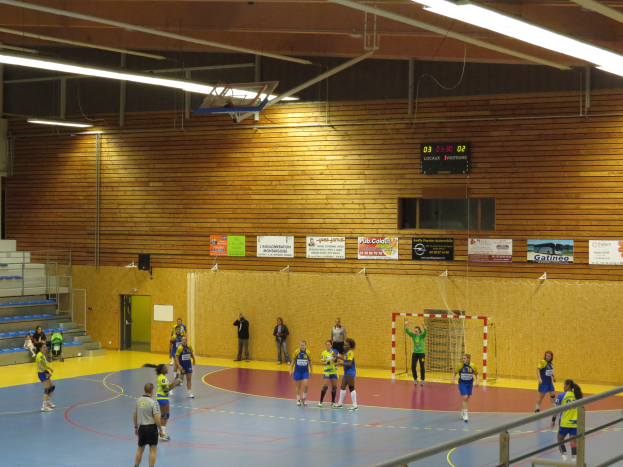 Eine Gruppe von Menschen, die Handball in einer Turnhalle mit einem Metallzaun, einem Tor, einer Tür, Texttafeln, einem Anzeigebildschirm, einem Lautsprecherkasten, Metallstangen und einem Dach mit Deckenleuchten und Metallrahmen spielt.