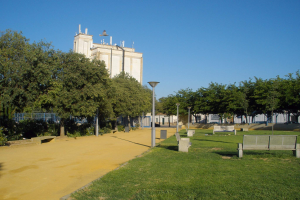 Ein Park mit Bänken und Bäumen vor einem Gebäude, umgeben von Straßenlaternen, Grills, Pflanzen und einem klaren blauen Himmel.