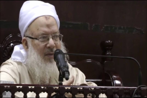 Asaduddin Owaisi, ein Mann mit einem weißen Bart und Turban, spricht in ein Mikrofon vor einer hölzernen Begrenzung, trägt ein cremefarbenes Gewand und Brille, vor einem dunklen Hintergrund.