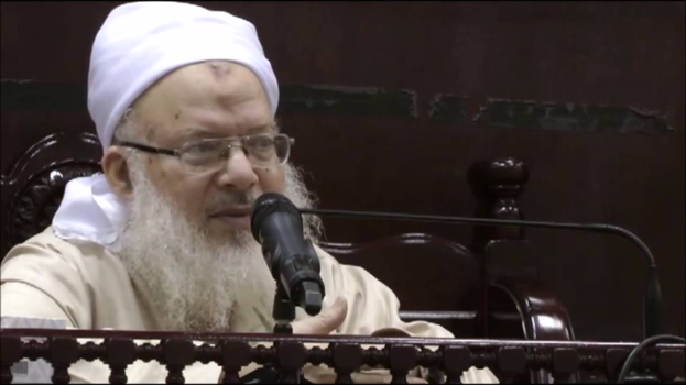 Asaduddin Owaisi, ein Mann mit einem weißen Bart und Turban, spricht in ein Mikrofon vor einer hölzernen Begrenzung, trägt ein cremefarbenes Gewand und Brille, vor einem dunklen Hintergrund.