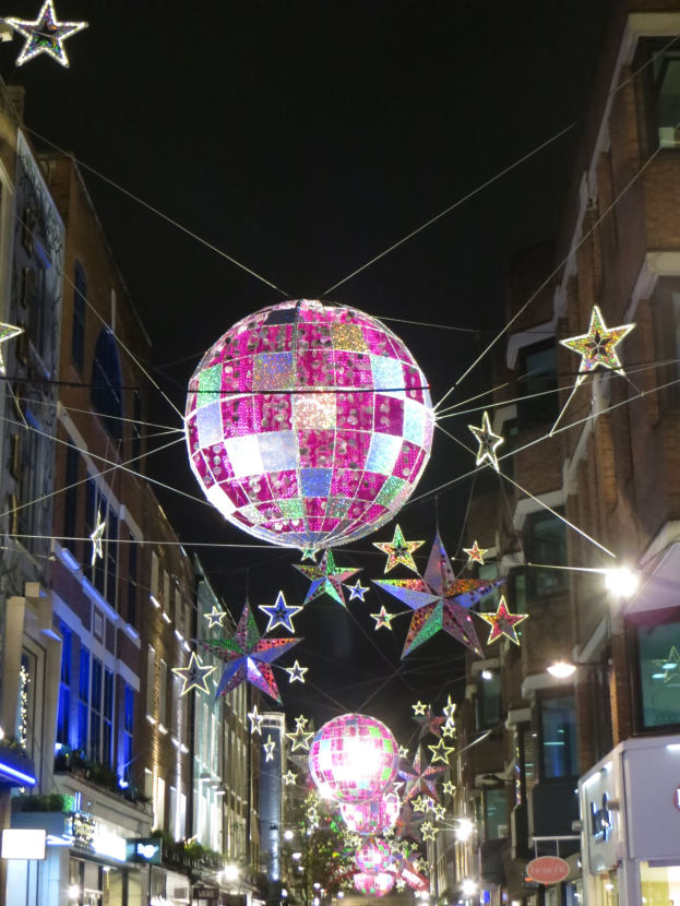 Eine Stadtstra├če bei Nacht mit zahlreichen Lichtern und Deko, mit einem gro├čen Diskokugel ├╝ber einem Geb├Ąude h├Ąngend, die eine festliche Atmosph├Ąre schafft.