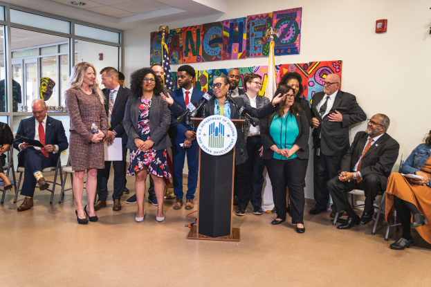 Eine Gruppe von Menschen, die in einem Raum stehen und sitzen, mit einem Rednerpult, einem Mikrofon und einem Schild mit der Aufschrift "Sancho-Stadtrat tritt dem Stadtrat bei", mit Flaggen, einer Tafel, einem Feuerlöscher und Deckenleuchten an der Rückwand.